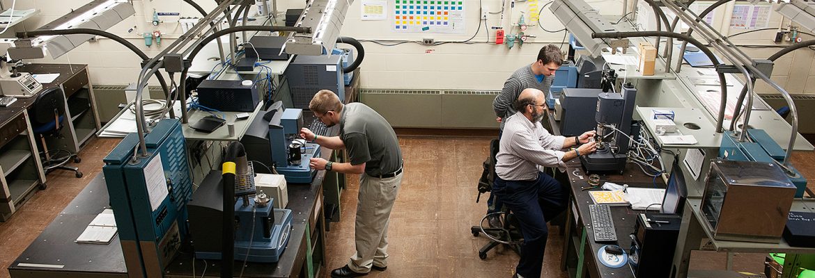 Three technicians operating precision fabrication and testing equipment in a laboratory workspace