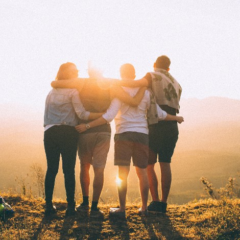 Group of four people with arms around each other looking at sunset on a mountain