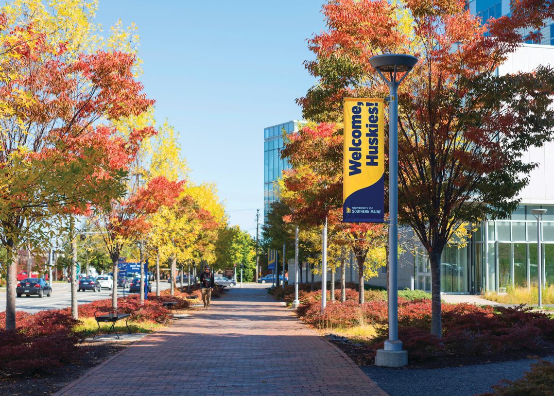Picture of walkway on Portland campus, the trees on the sides are turning red for the fall. A "welcome huskies" flag hangs from a lamp post.