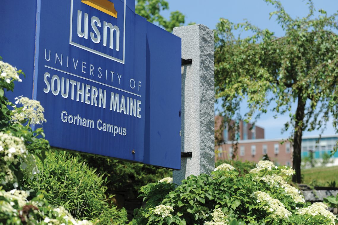 "University of Southern Maine" welcome sign for the Gorham Campus. It is dark blue with white text, surrounding by green plants and white flowers.