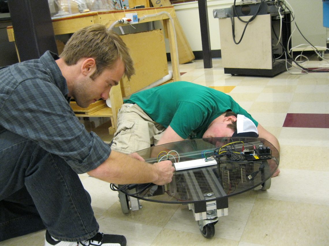 Students kneeling on a laboratory floor adjusting the wires of a device that consists of a circular piece of dark-colored glass or clear plastic with a T-shaped support apparatus beneath it on wheels.