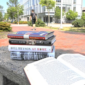 A stack of books including "The Prince of Los Cocuyos," "American Creation," "At Home," "Born to Run," and "The Complete Sherlock Holmes" rest on a bench in front of the Wishcamper Center.