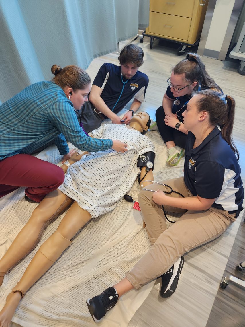 A patient mannequin lies on its back on the floor of a medical examination room. FOur students sit around it, checking its vitals with medical equipment.