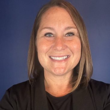 A photo of Amanda Eisenhart; a woman with light brown hair stands in front of a blue background smiling.