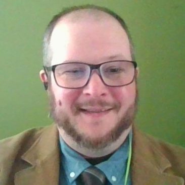 A photo of Benjamin Jones; a man with a beard stands in front of a green background smiling.