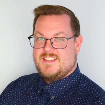Headshot of Chandler Guptill. A man smiling at the camera; he is wearing glasses and a blue collared shirt with white dots.