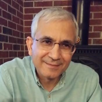 Headshot of a man with short white hair and a green shirt and glasses in front of a brick wall.