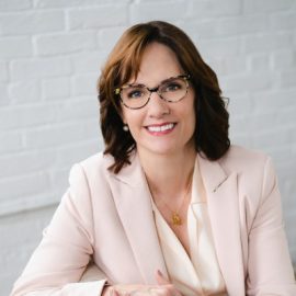 Lisa Walker smiling in a light pink blazer with a white painted brick background