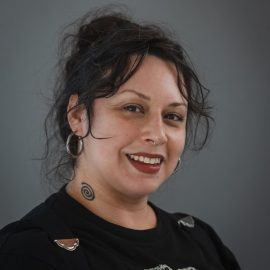 portrait of person with long dark brown hair and bangs looking at the camera. They are wearing a light blue button down shirt with a collar. The background is blurry greenery