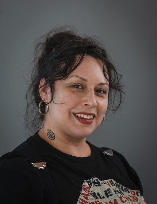 portrait of person with long dark brown hair and bangs looking at the camera. They are wearing a light blue button down shirt with a collar. The background is blurry greenery