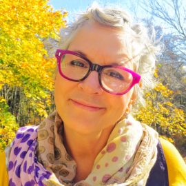 Photo of Jennifer Bragdon. She is wearing a multi-colored polka dot scarf, with purple glasses, short wavy grey hair, light skin, and brown eyes. She is standing in front of a bright green tree and is smiling.