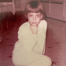 This is a photograph of Marcia taken when she was 8 years old. She is wearing white footie-pajamas and sitting on her 1960s linoleum kitchen floor.