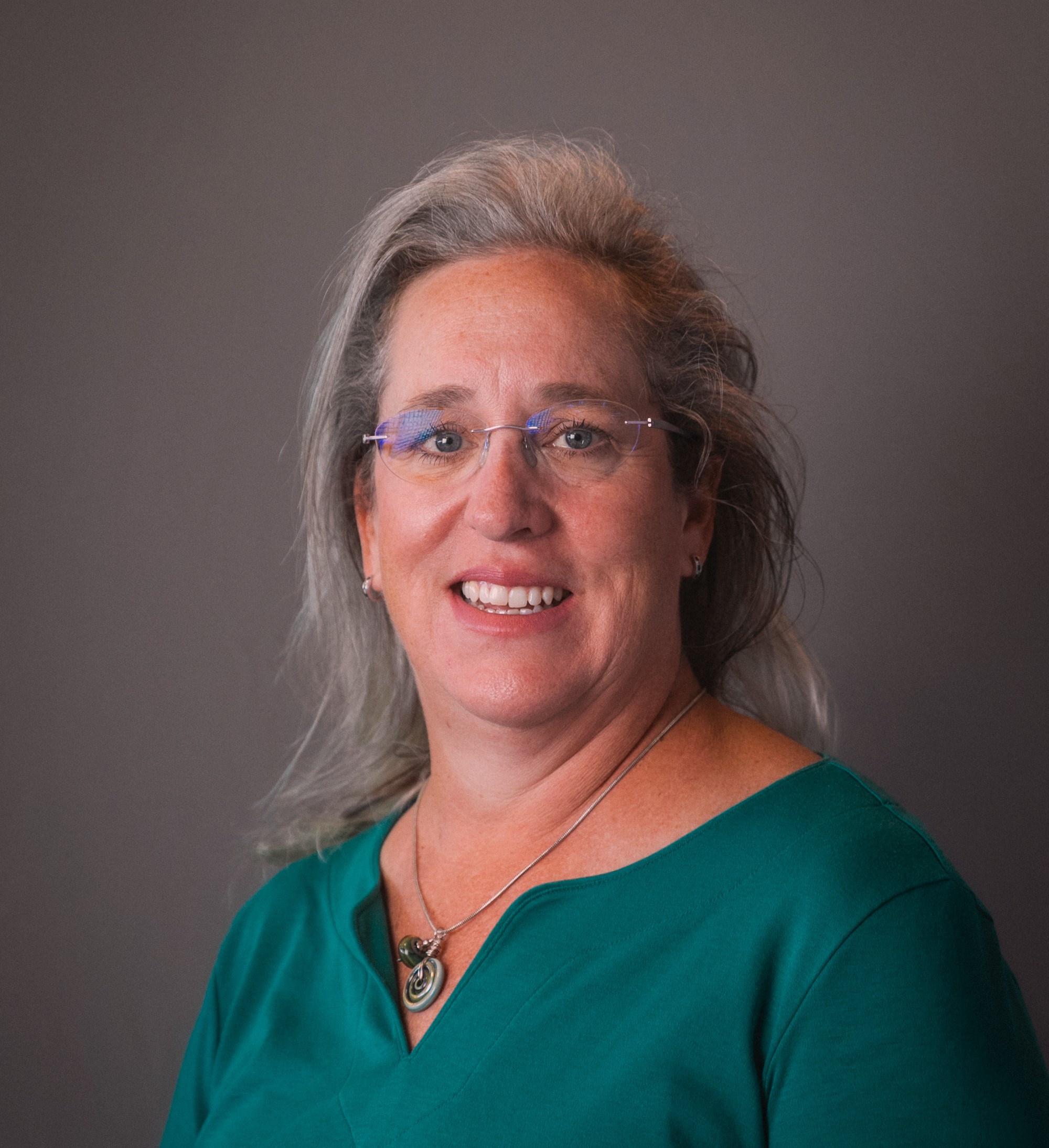 A headshot of Leslie. She has glasses, long hair, a green top, and is smiling.