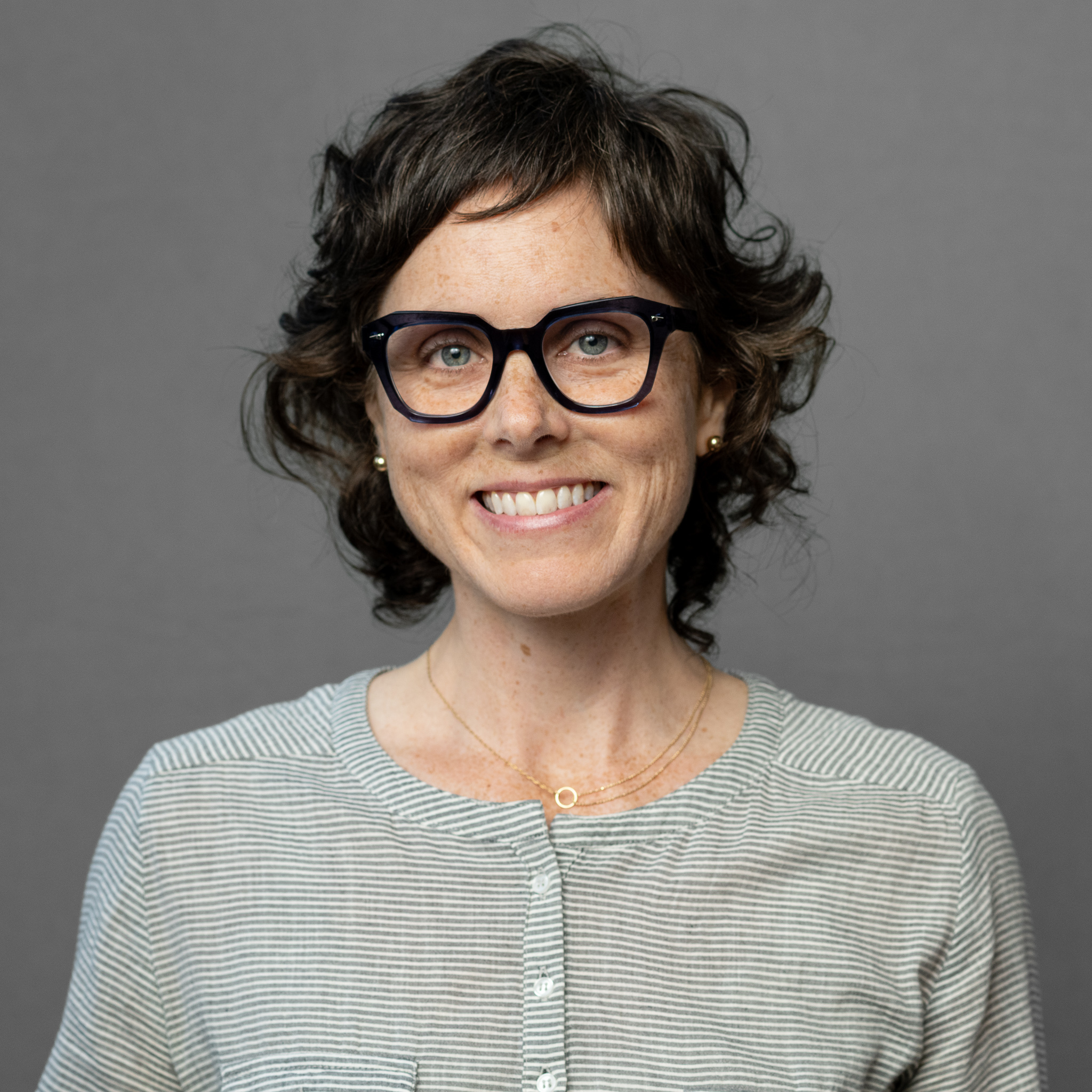white woman with short dark brown hair, dark blue thick rimmed glasses, and gray shirt smiling