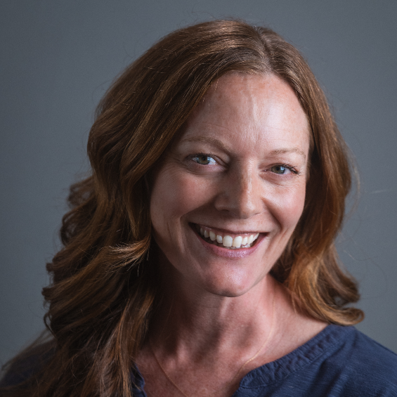 A smiling woman with reddish hair wearing a blue top in front of a gray portrait background.
