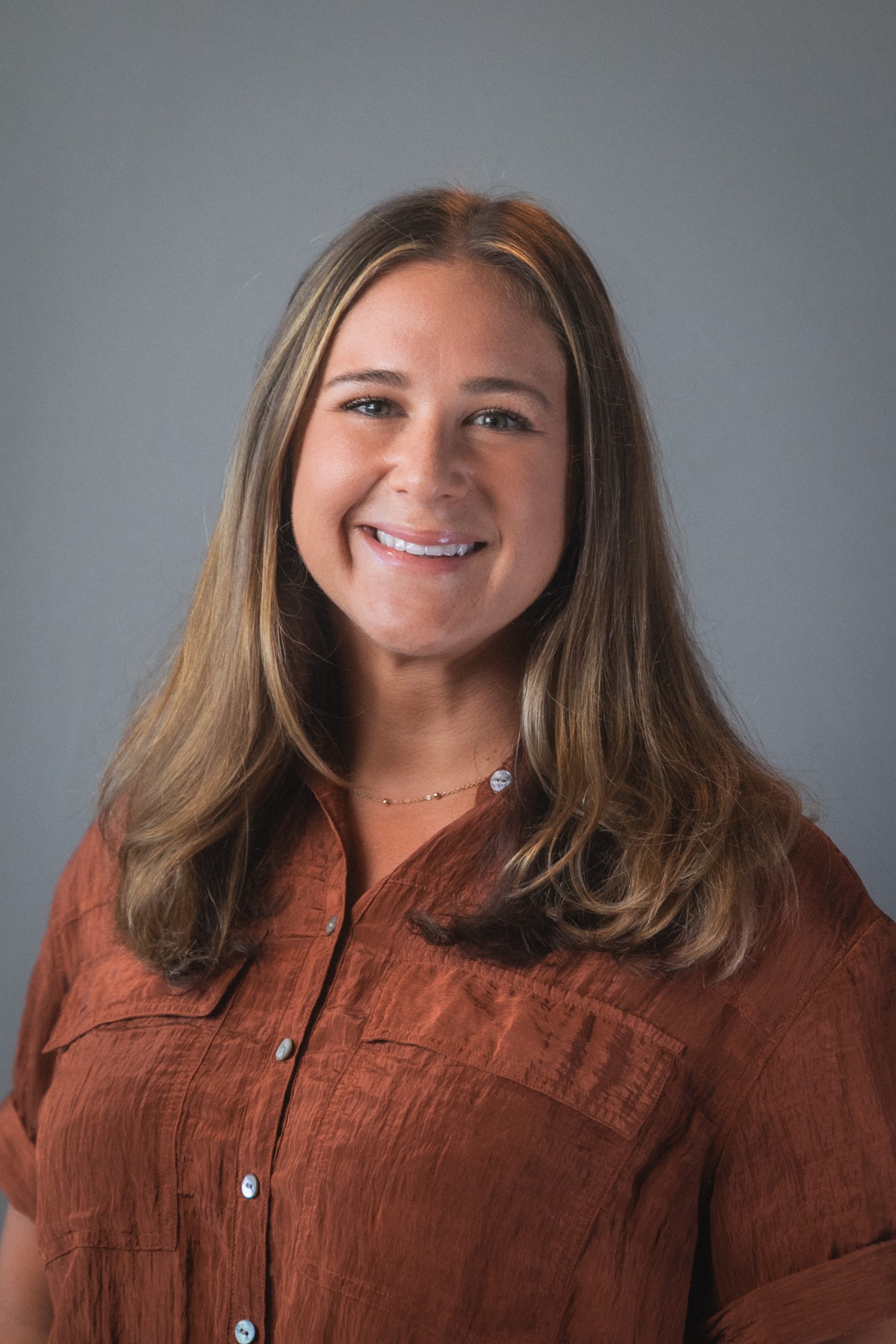 Suzanne has long brown hair and is smiling and is wearing a brown shirt.