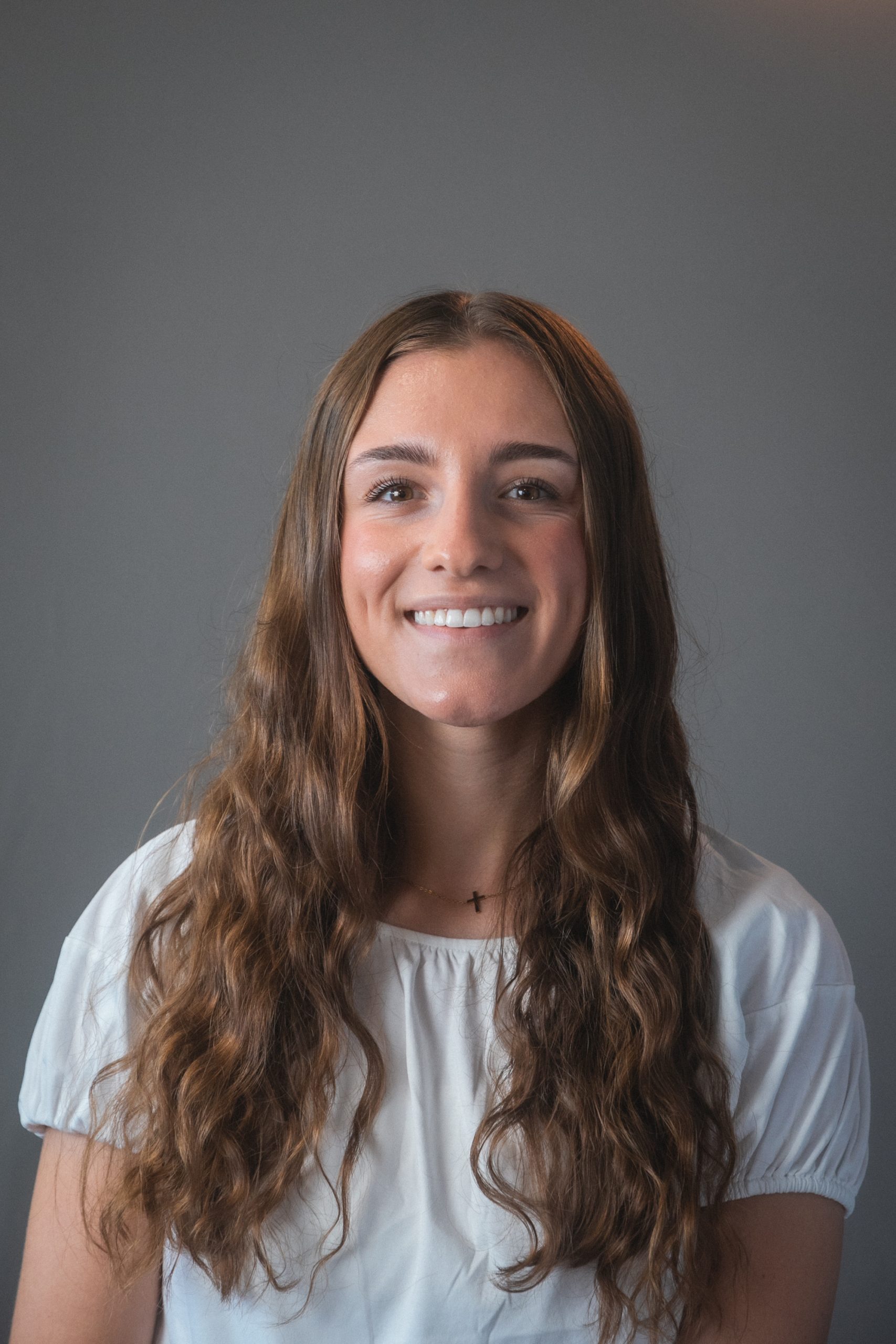 A smiling woman with long wavy brown hair and a white top in front of a gray background