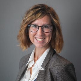 A photo of Christina. She has glasses, chin length hair, and is smiling. She is wearing a blazer and white shirt.