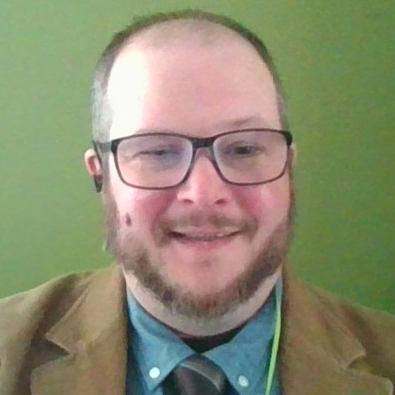 A photo of Benjamin Jones; a man with a beard stands in front of a green background smiling.