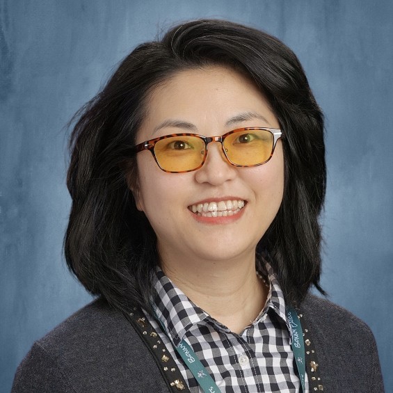 A picture of Sherry Hu; a dark-haired woman standing in front of a blue background and smiling. She's wearing turtle-brown glasses with an orange tint.