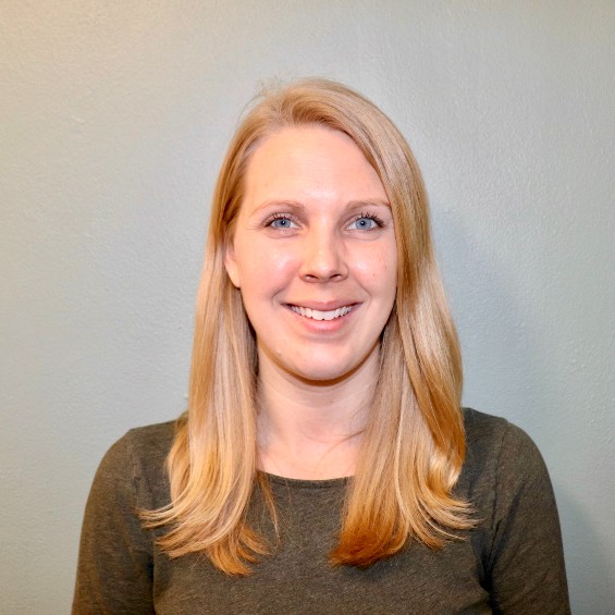 A picture of Victoria Petersen; a blonde woman standing in front of a gray background and smiling.