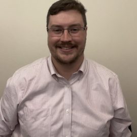 Tim is wearing glasses and a striped shirt and is smiling.