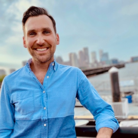 A smiling man with dark hair wearing a blue shirt standing on a bridge in front of a large city skyline.