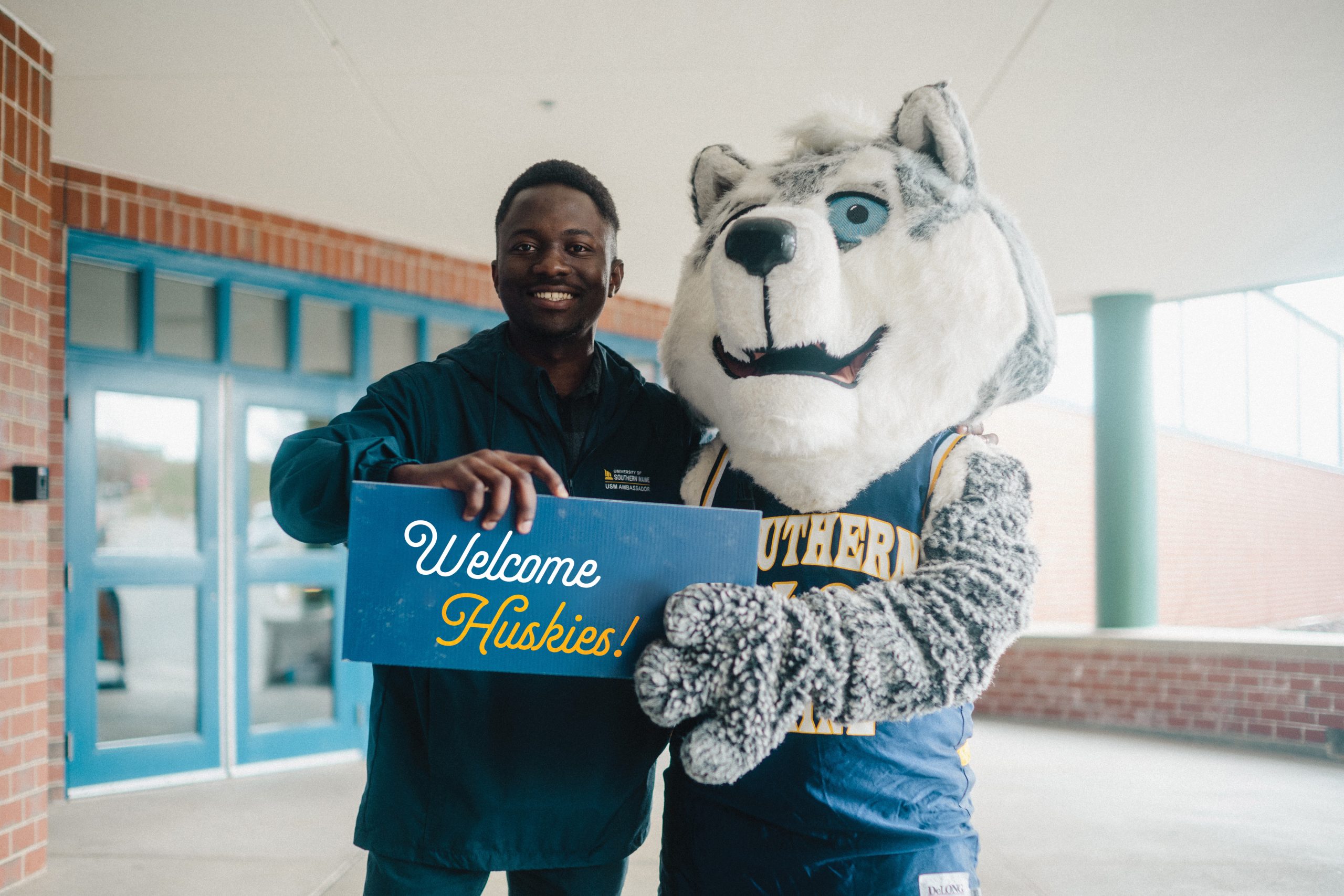 Student stands next to Champ mascot, holding 'Welcome Huskies' sign.