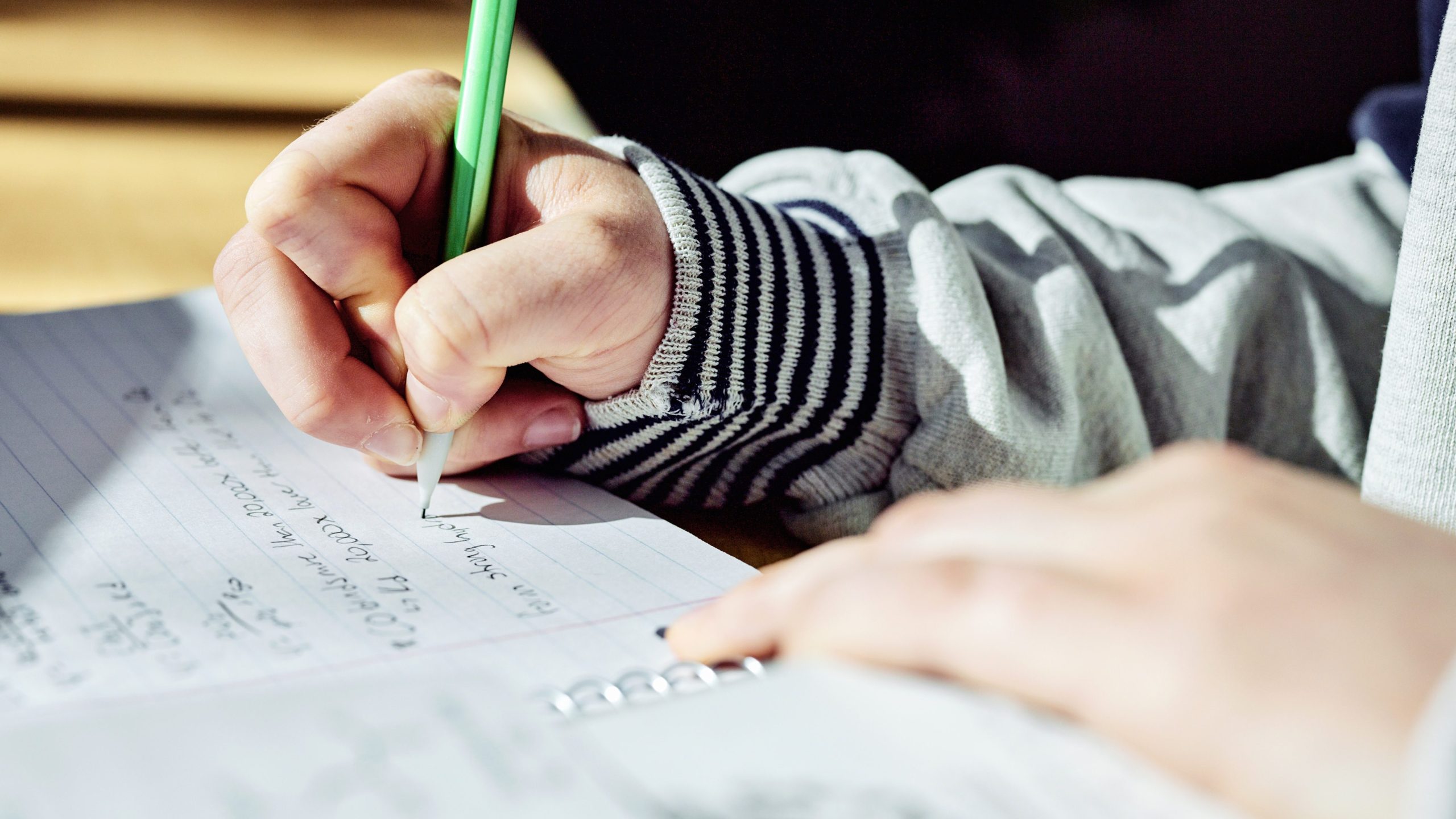 Zoom in on student gripping green pencil, writing in notebook.