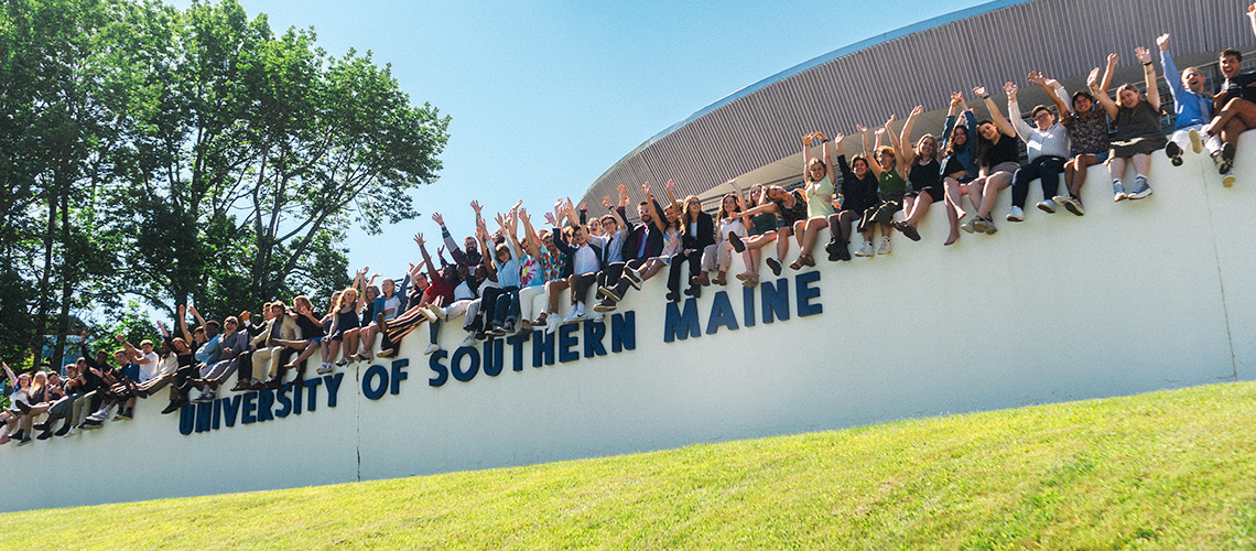 A large group of students sit with their arms raised in the air, on top of a wall that spells out 'University of Southern Maine".