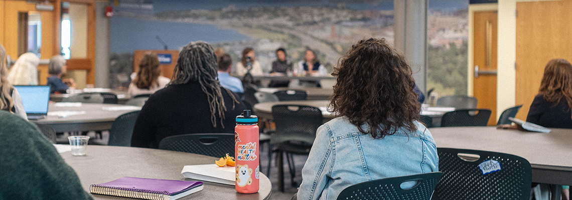 Student sit and listen to a panel discussion, one with a water bottle featuring a sticker stating "mental health matters".