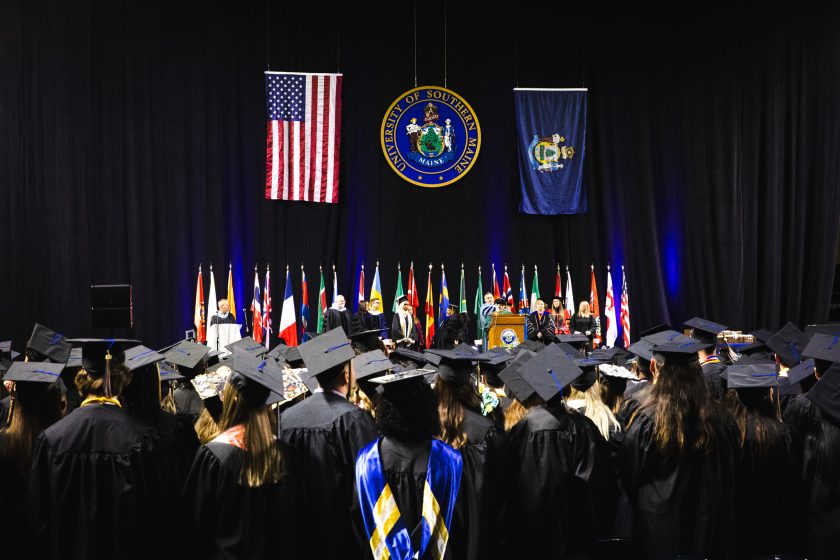 Graduates in black caps and gowns face the stage at Cross Insurance Arena during USM's 146th Commencement ceremony, with the University seal, American flag, and Maine state flag displayed above the stage party.