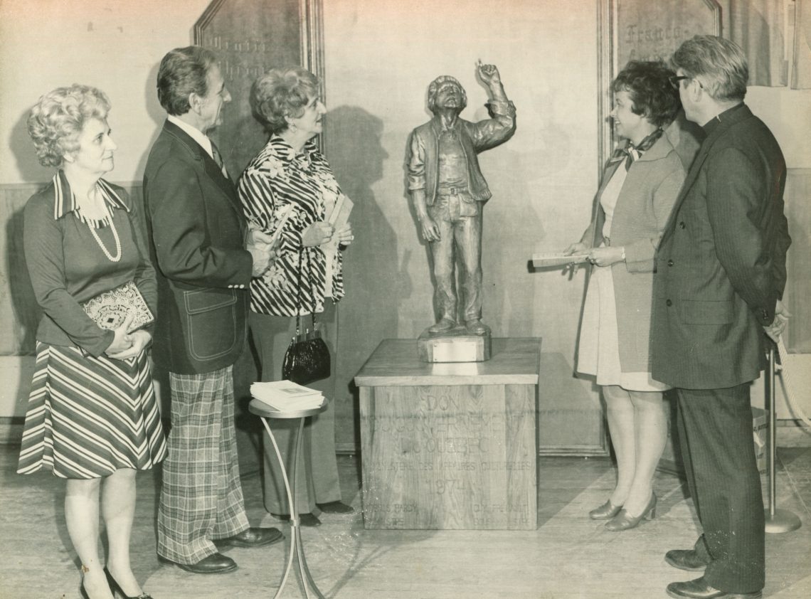 Lewiston-Auburn dignitaries stand around the Paysan Quebecois wooden statue carved by J.J. Bourgault in Lewiston in the 1970s. The statue is three feet tall male figure, holding his figure up and in mid-speech.
