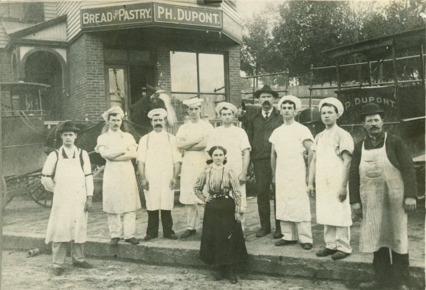 Image of bakers in front of Dupont Bakery in Auburn in 1893. 9 men and one woman pose in front of the brick bakery building next to delivery carriages and horses. Seven of the men are wearing bakers hats and white aprons. One of the men, the owner, is wearing a suit. 
