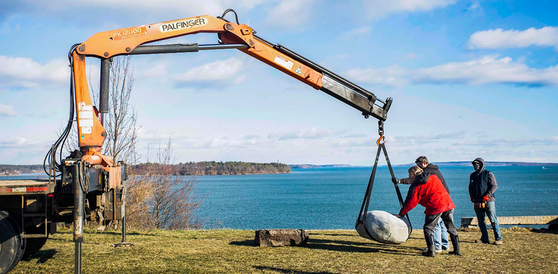 A construction crane drops a large boulder onto a vista overlooking the ocean.