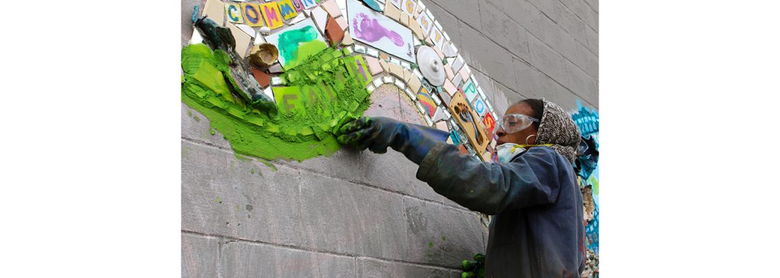 A woman with a mask creates a mosaic on a cinderblock wall.