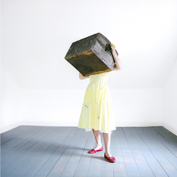 An image of a woman in a room wiht white walls and a grey floor. She wears red flats and a yellow dress. On her head is a large, rectangular box, obscuring her head and neck. Her hands reach up onto the box's sides; she cannot reach its top.