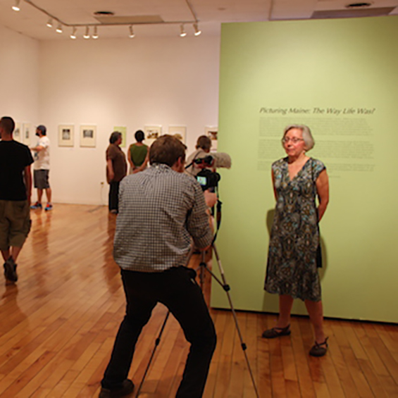 An image of a gallery with wooden floors and white walls. In the froreground a photographer with a tripod has his back to the viewer, facing him is a woman in a dress standing in front of a green wall.
