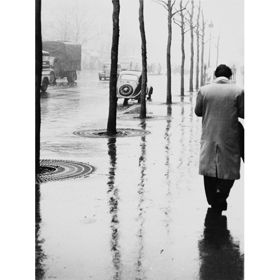 A black and white photograph of a man walking, wearing a trench coat. His back is to the viewer. It is raining and there are reflections of the man at right and reflections of lamposts at left.