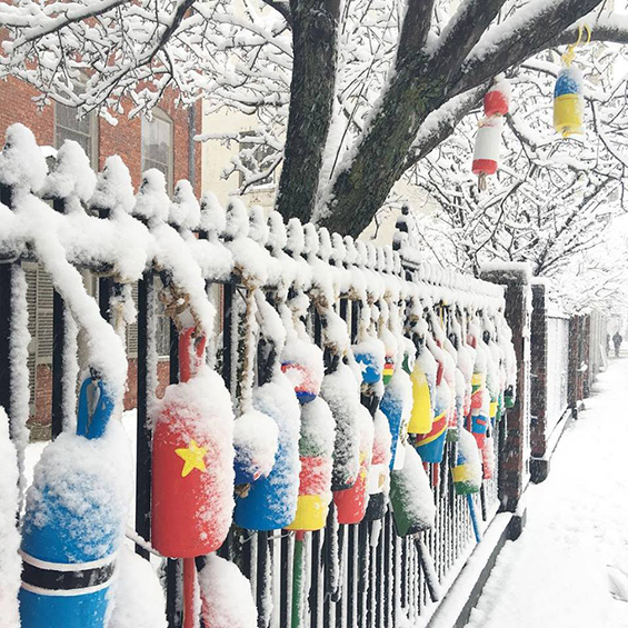 An image of buoys hanging on an iron fence. Each buoy is painted like the flag of a different country. There is snow on the fence, ground, and buoys.