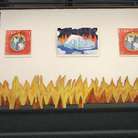 An image of a gallery. Three drawings are on the wall. In the foreground are cardboard cut outs of flames