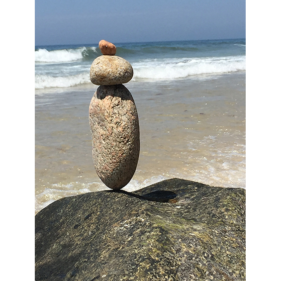 A photograph of three small stones balanced atop one another. They rest on a large boulder that goes out of the bottom of the frame. In the background is a beach with waves.