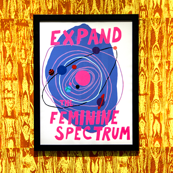 An image of a print hanging on a wall. The wall has drawn wooden wallpaper. The print has a series of planetary-like designs and their orbit. It reads "EXPAND THE FEMININE SPECTRUM."