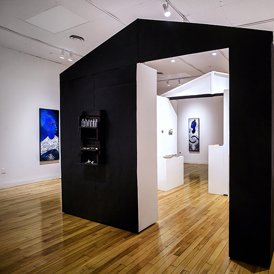 An image of a gallery with primarily blue-palette portraits on the walls. A structure resembling a house, painted black, sits in the foreground. Its left wall reads "OTHERED"