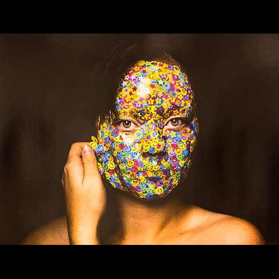 An image of a woman whosse face is covered in smile stickers of different colors. They are overlapping and form one sheet. She is beginning to peel the sheet off from the left-hand side.