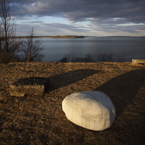An image of two rocks overlooking a large body of water. One is a light grey on the right; the other is more angular and black on the left. They cast long shadows on grass pointing upwards and right. The sky above is stormy.