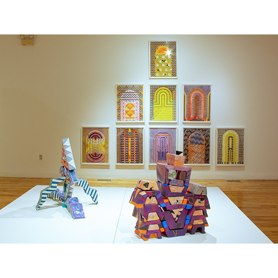 A Gallery with abstract sculptures in the foreground and obelisk watercolors arranged in a pyramid on a wall in the background