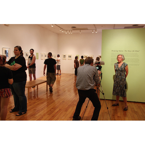 An image of a gallery with wooden floors and white walls. In the froreground a photographer with a tripod has his back to the viewer, facing him is a woman in a dress standing in front of a green wall.