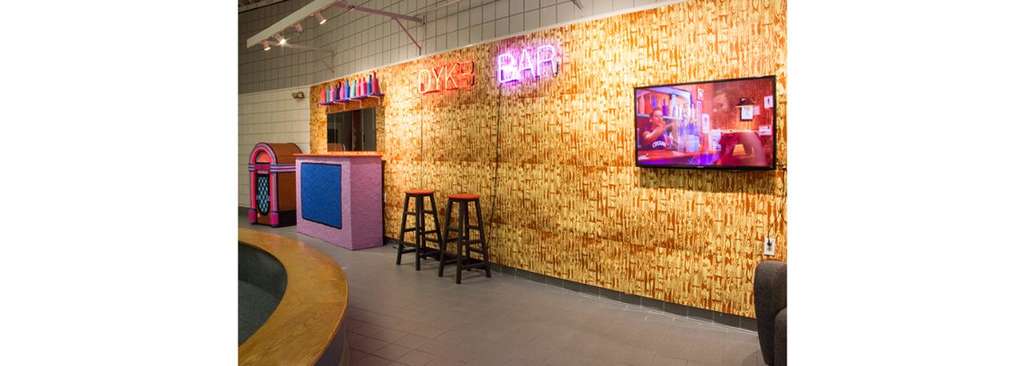 An image of a room with a neon sign that reads "DYKE BAR." There are barstools and the walls are covered in a faux wood grain wallpaper.