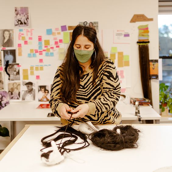 An artist is braiding synthetic hair at a table. The artist has long hair and is wearing a tiger print long sleeve top and face mask. They have long hair and there are post its on a white wall behind them.